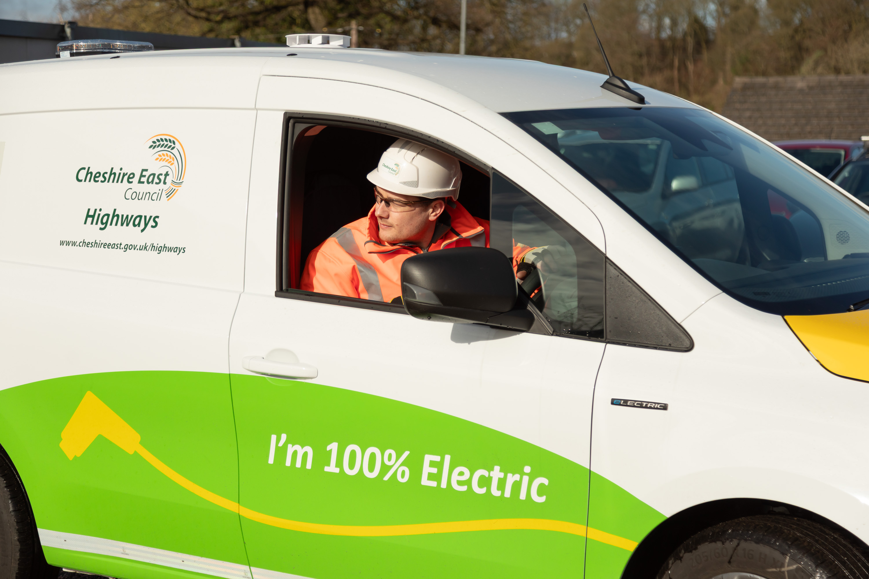 Highway worker wearing a hard hat and highvisibility jacket driving a white electric service van marked “I’m 100% Electric” for Cheshire East Highways, used by Ringway Jacobs as part of its carbon reduction strategy.